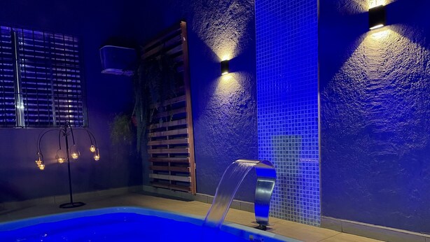 Ein Schwimmbecken mit Wasserspiel, umgeben von Wänden mit blauen, strukturierten Fliesen und einer Holzlamellentür.