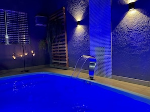 Ein Schwimmbecken mit Wasserspiel, umgeben von Wänden mit blauen, strukturierten Fliesen und einer Holzlamellentür.