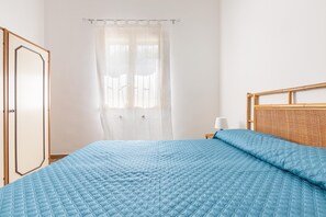 Ein Schlafzimmer mit einem Bett, einem hölzernen Kopfteil, einer weißen Tür und einem Fenster mit durchsichtigen Vorhängen.
