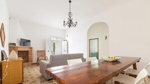 A dining area with a wooden table, white chairs, a grey sofa, a wicker cabinet, and a television on a stand. - Villa Bella Giulia by BarbarHouse (Maruggio)