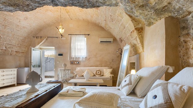 Ein gemütliches Schlafzimmer mit Steindecke, einem Bett mit weißer Bettwäsche, einem kleinen Tisch und einem Fenster mit Vorhängen.