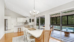 Een moderne keuken met witte kasten, een eethoek met een witte tafel en rieten stoelen, en een grote schuifpui naar een zwembad.