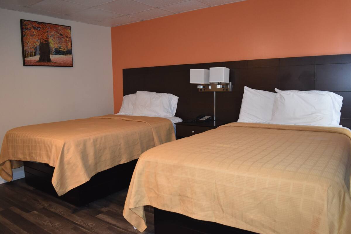 Two beds with beige bedding, a nightstand with a lamp, and a painting on the wall.