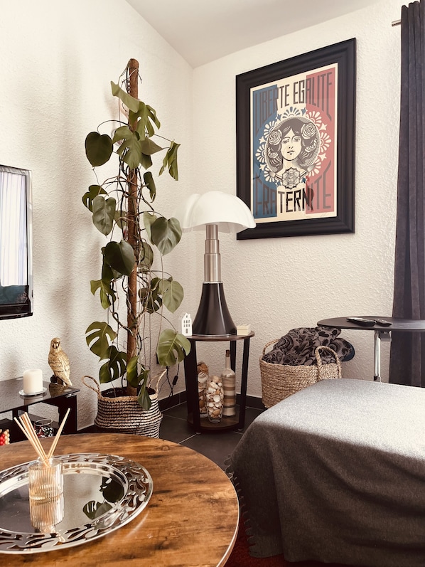 Ein modernes Hotelzimmer mit einer großen Kübelpflanze, einem runden Holztisch mit Tablett, einer schwarzen Lampe, einem gerahmten Poster und einem Bett mit grauer Decke.