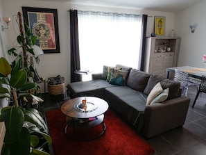 Ein modernes Wohnzimmer mit einem grauen Sofa, einem roten Teppich und einem Couchtisch. Ein Fenster mit Vorhängen, ein kleiner Tisch und ein Regal mit dekorativen Gegenständen sind ebenfalls vorhanden.