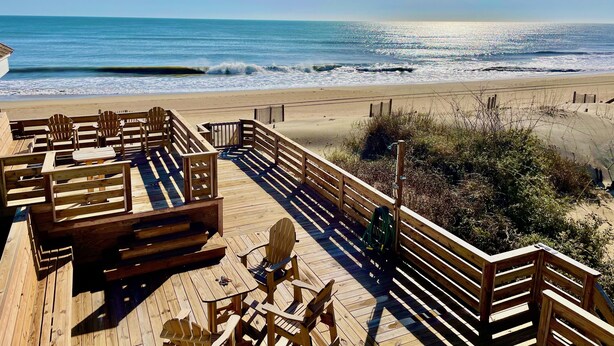 Une terrasse en bois, agrémentée de chaises et de tables de style Adirondack, donnant sur une plage où l’on peut admirer les vagues.