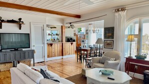 Un salon spacieux avec une grande télévision, des meubles en bois, un coin repas et une cuisine à l’arrière-plan.