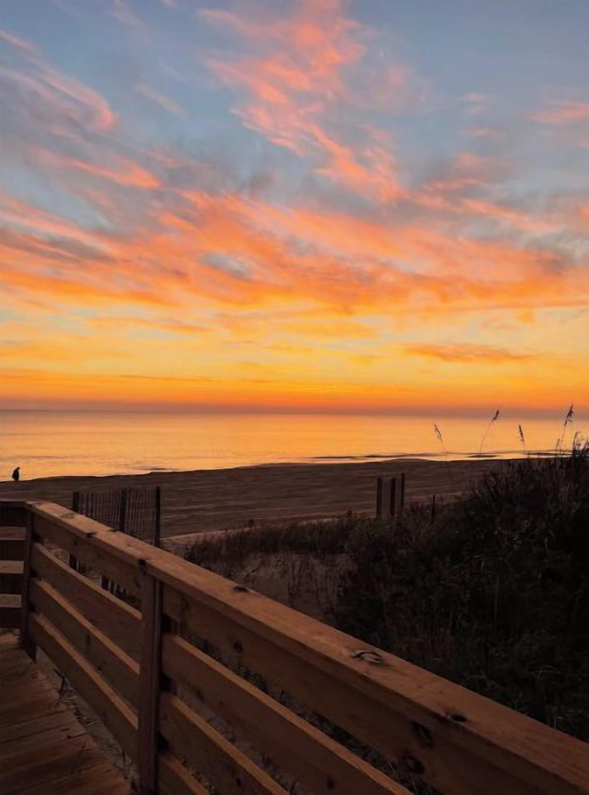 Terraza de madera con vista a una playa al atardecer, bajo un cielo despejado.