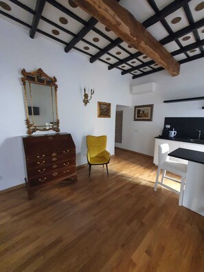 Quarto com piso de madeira, uma cadeira amarela, uma cômoda de madeira com gavetas, um espelho grande e uma escrivaninha branca com bancada preta.