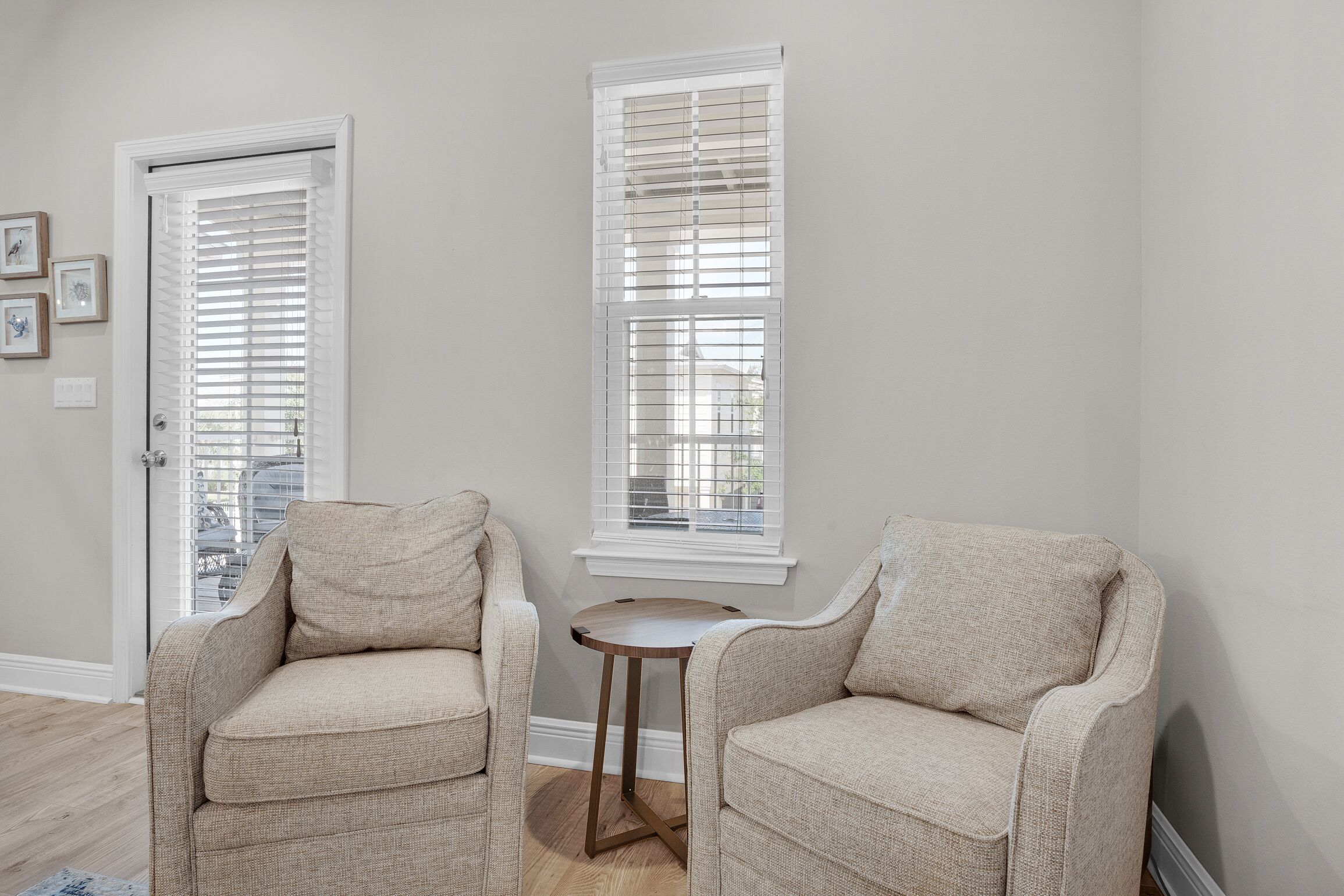 A room with two beige armchairs, a small round table, and two windows with blinds.
