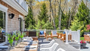 Um deck de madeira com mobiliário para áreas externas, guarda-corpo branco e vista para árvores e um edifício.