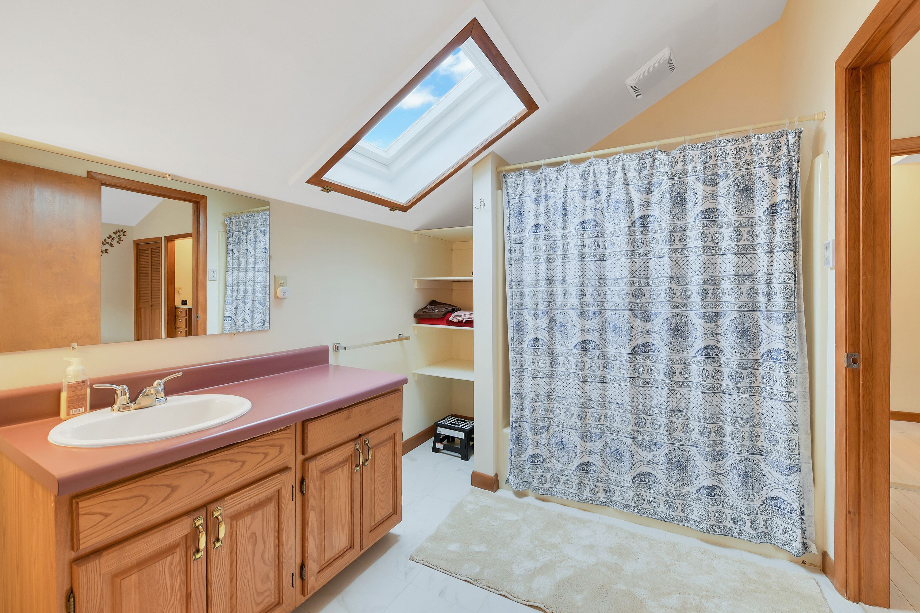A bathroom with a skylight, a sink, wooden cabinets, and a patterned shower curtain.
