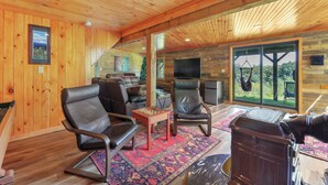 A cozy living room with wooden walls and ceiling, a large window, a fireplace, and a television.