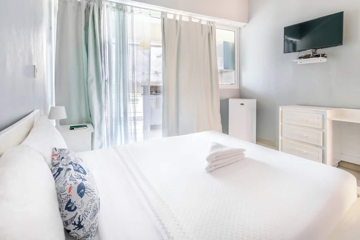 A neatly made bed with a white comforter and a decorative pillow, a bedside table with a lamp, a TV mounted on the wall, and a view of a balcony through a window with curtains.