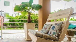 A wooden lounge chair with patterned cushions, a potted plant, and a view of greenery and a building in the background. - Bella Vida Hotel Punta Cana (Punta Cana)