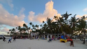 A beach with palm trees, a shopping center sign, and beach chairs. - Bella Vida Hotel Punta Cana (Punta Cana)