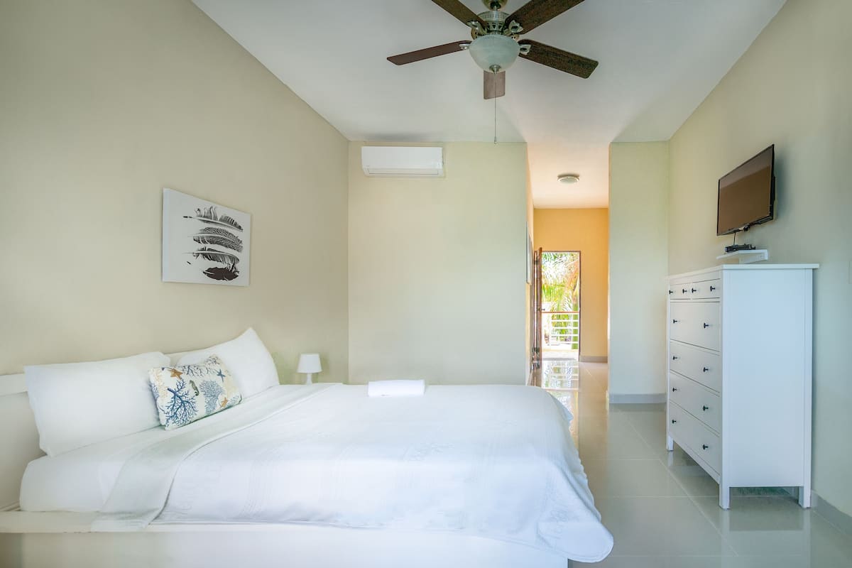 A bedroom with a bed, a dresser, a ceiling fan, a TV, and a window with a view.