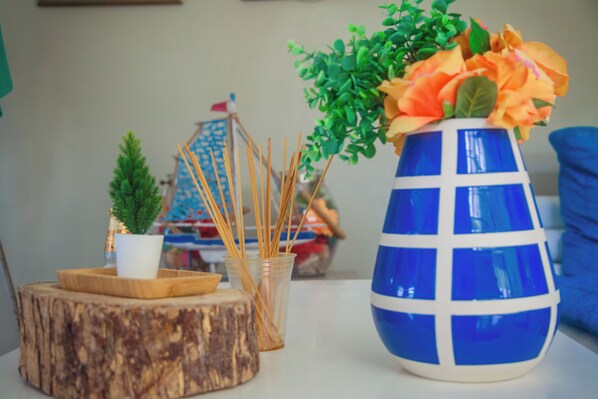 Una mesa con un jarrón azul con flores naranjas, una planta pequeña en maceta y un recipiente de vidrio con palitos de madera.