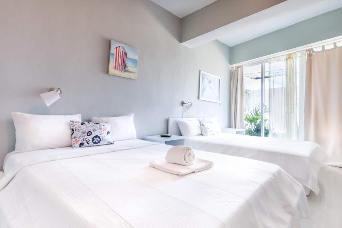 A hotel room with two beds, a nightstand, a lamp, and a window with curtains.