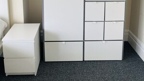 A white wardrobe with multiple compartments and drawers, a white bedside table, and a dark carpeted floor. - Abbeyfield Guesthouse (Liverpool)
