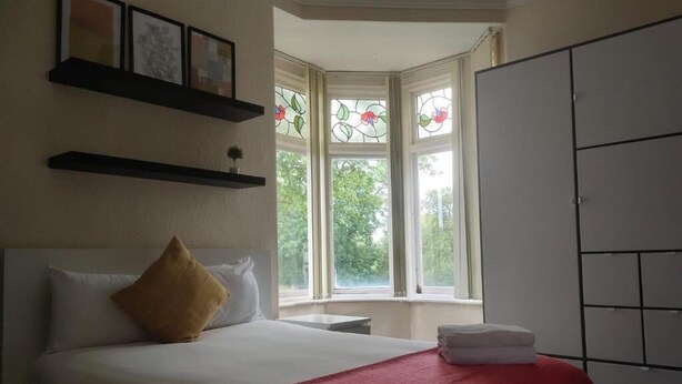A hotel room with a bed, a red bedspread, a window with stained glass, and a wardrobe. - Abbeyfield Guesthouse (Liverpool)