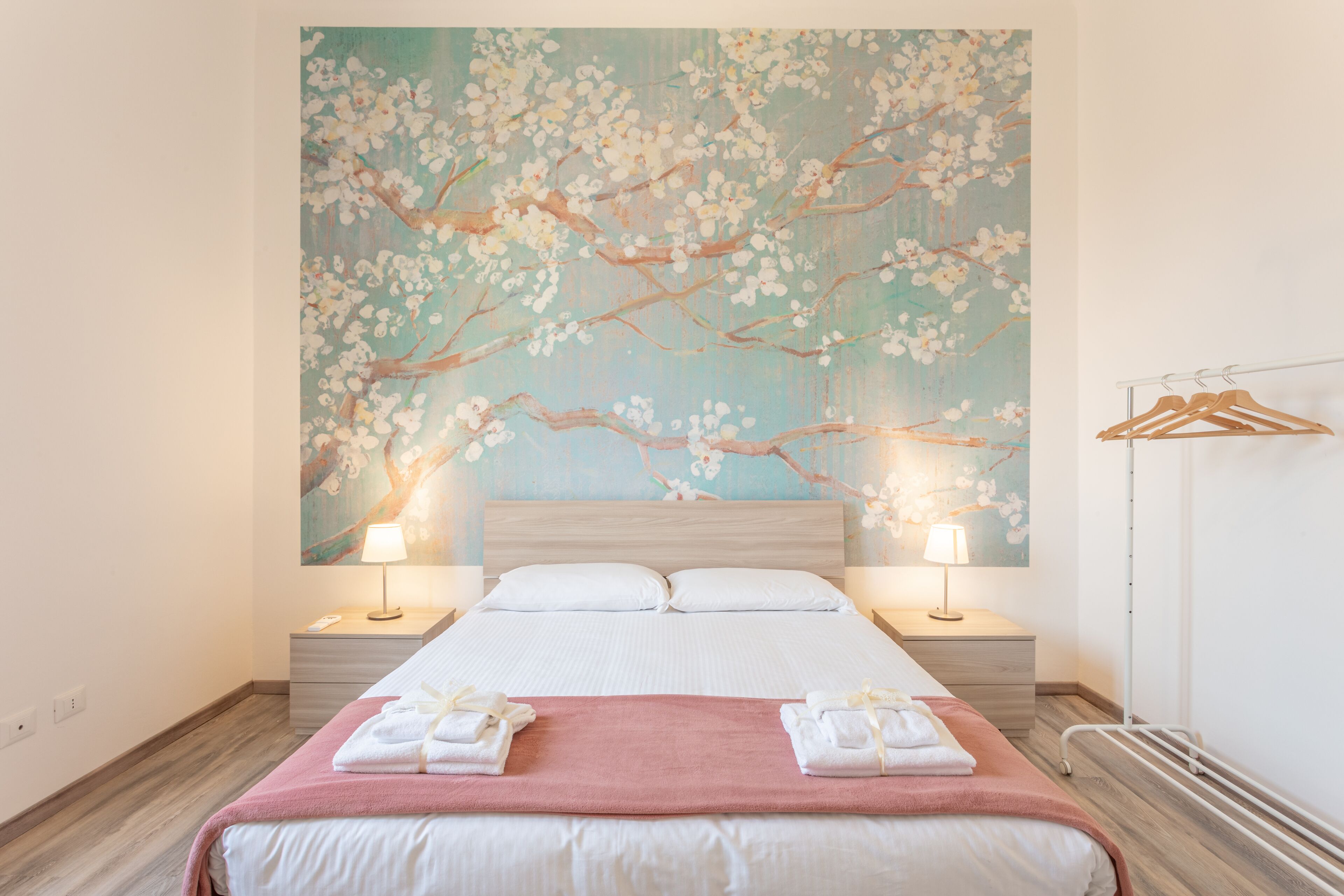 A hotel room with a large floral mural on the wall, a neatly made bed with a pink blanket, two bedside tables with lamps, and a metal radiator.
