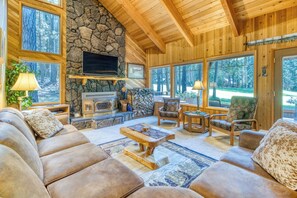 Un interior acogedor con chimenea de piedra, techo de madera y ventanas amplias que dan a un bosque.