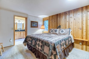 Quarto com parede de madeira, cama com colcha estampada, mesa de cabeceira com abajur e banheiro visível por uma porta aberta.