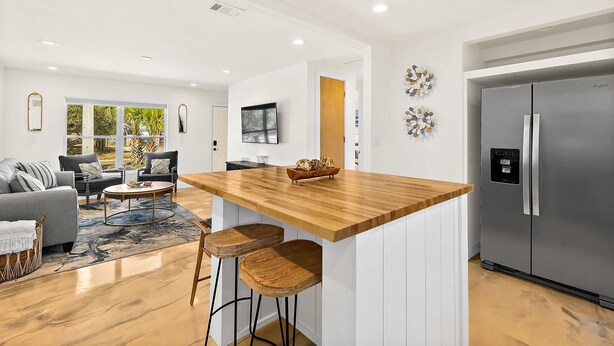 Una cocina moderna con una isla central, taburetes de bar, un refrigerador grande y una zona de comedor con sofá y mesa de centro.