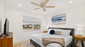 Un dormitorio con cama, ventilador de techo, ventana con vistas, un cuadro y televisor sobre un soporte.