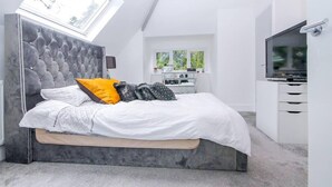 A modern bedroom with a tufted headboard, a bed with white and gray bedding, a bright window, and a TV on a white cabinet. - Luxury 3-bed Penthouse Apartment in Bournemouth (Bournemouth)