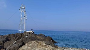 Una costa rocosa con un bote blanco y una alta estructura metálica, posiblemente un faro o torre de comunicaciones, frente a un cielo azul despejado.