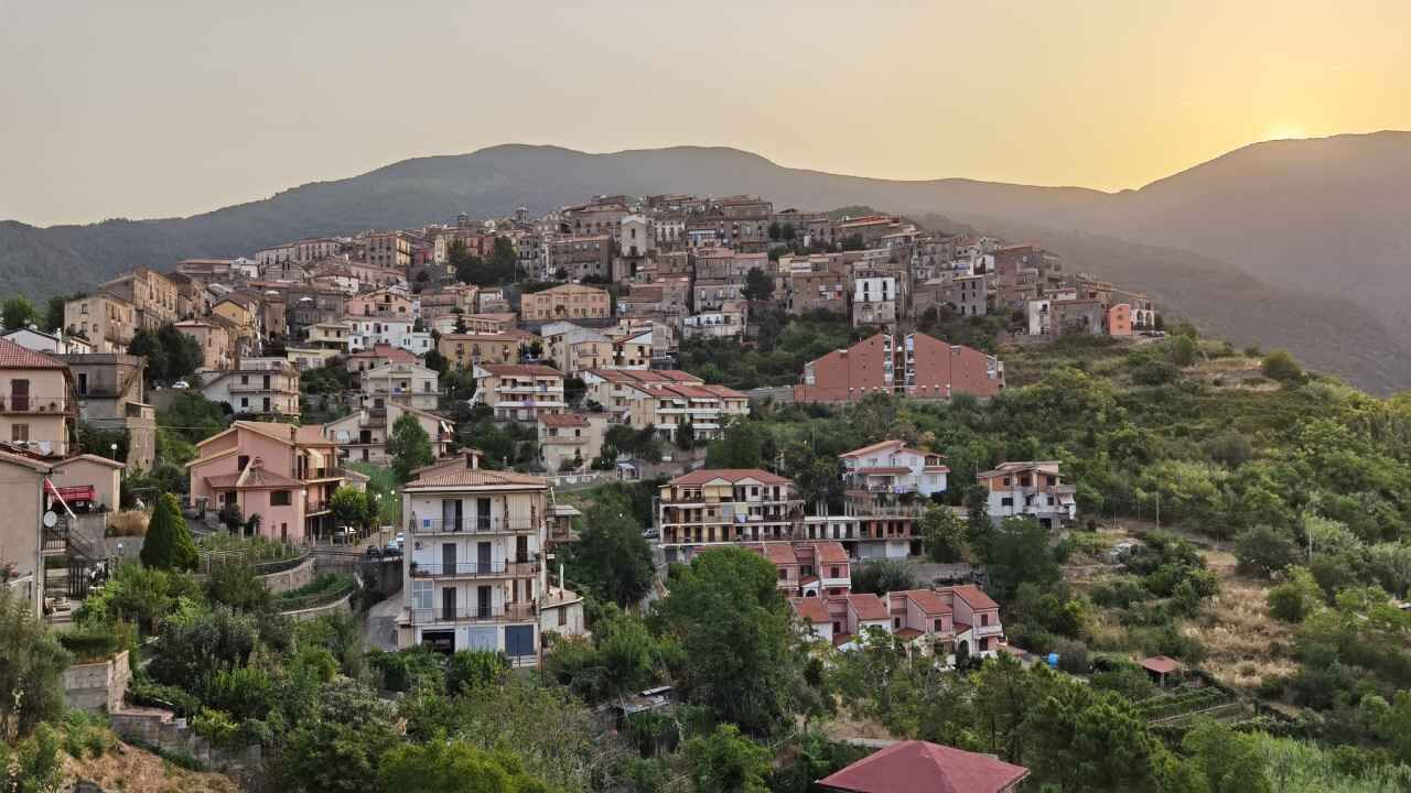 A hillside town with buildings clustered together, surrounded by greenery and mountains in the background.