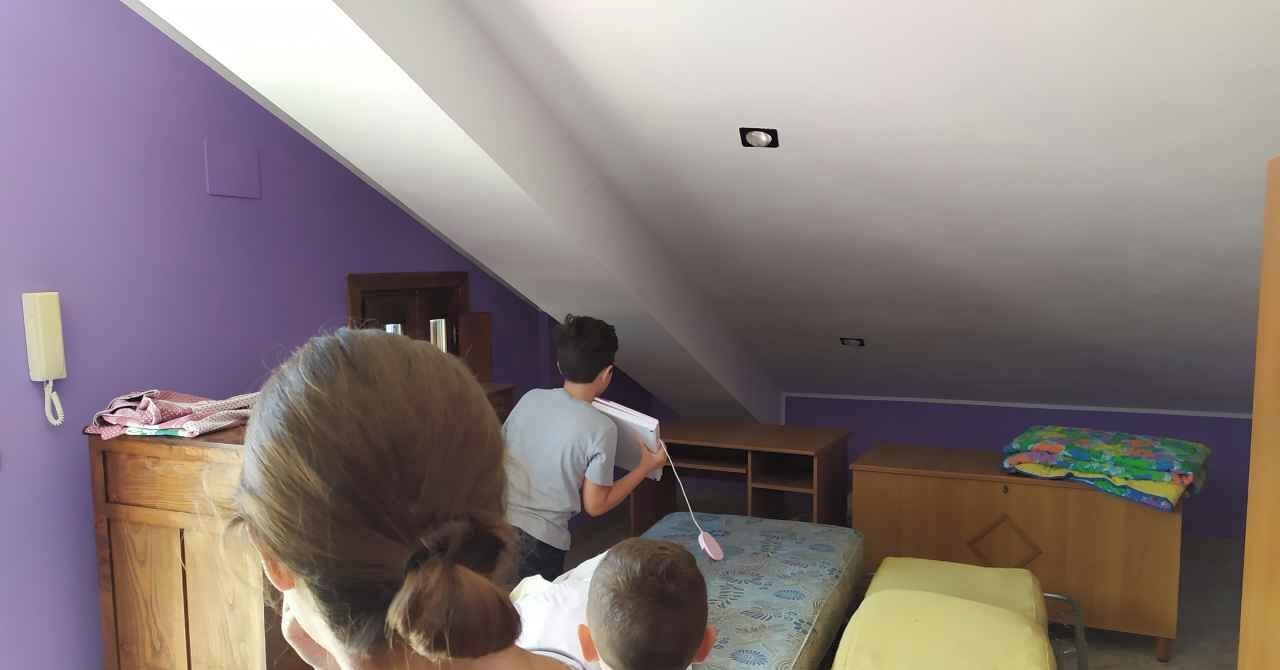 Une pièce aux murs violets, avec un bureau en bois, un lit orné d’un coussin jaune, et un enfant qui joue avec un jouet.