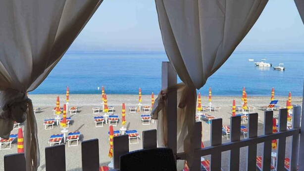 Vista de la playa con hileras de sillas y sombrillas, una barandilla y el mar al fondo.