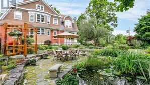 A two-story house with a brick facade, a wooden porch with a swing, and a garden with a pond and various plants. - Eddie Manor: 1907 Gem in Downtown Meaford (Meaford)