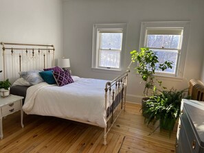 A bedroom with a metal bed frame, a white bedside table, a nightstand, a dresser, two windows, and a potted plant. - Eddie Manor: 1907 Gem in Downtown Meaford (Meaford)