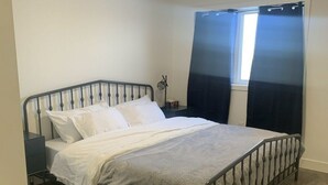Een enkel bed met een metalen frame, wit beddengoed en een grijze deken. Er is een klein nachtkastje met een lamp, een raam met donkere gordijnen en een houten vloer.