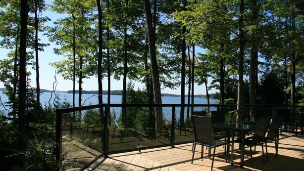 A wooden deck with a glass railing overlooking a lake, surrounded by tall trees. - Lake Eugenia Autumn Escape - Private Dock, Hot tub and Firepit (Flesherton)
