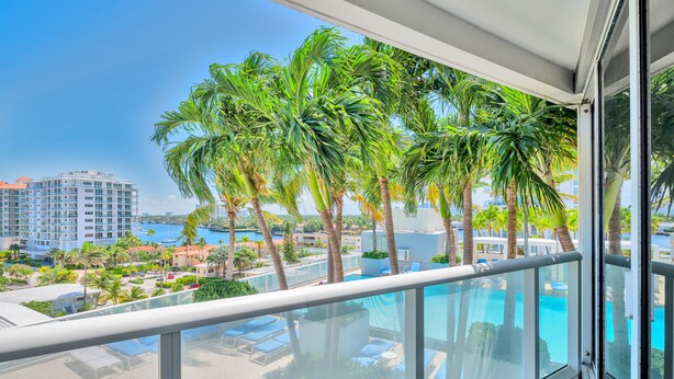 A balcony with a glass railing overlooking a swimming pool, palm trees, and a cityscape. - W Fort Lauderdale Residences (Fort Lauderdale)