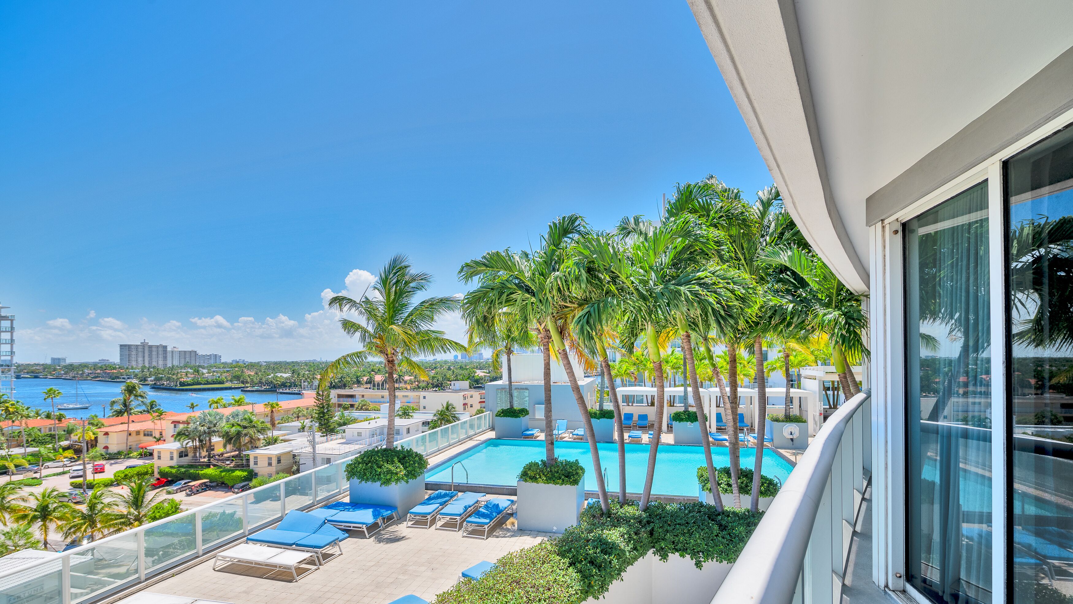 A rooftop pool area with palm trees, lounge chairs, and a view of the ocean and city skyline.