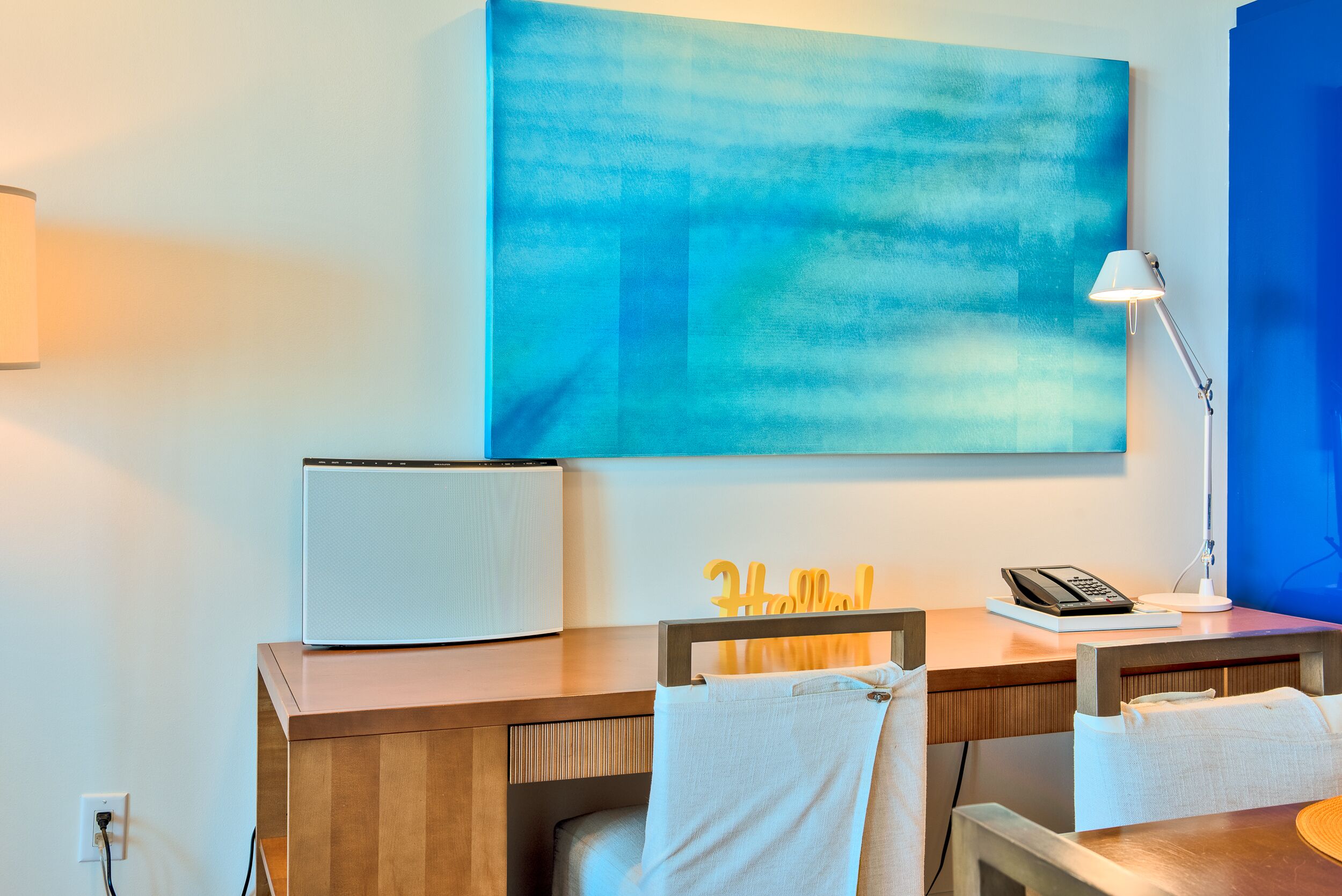A hotel room with a wooden desk, a laptop, a telephone, and a large abstract painting on the wall.
