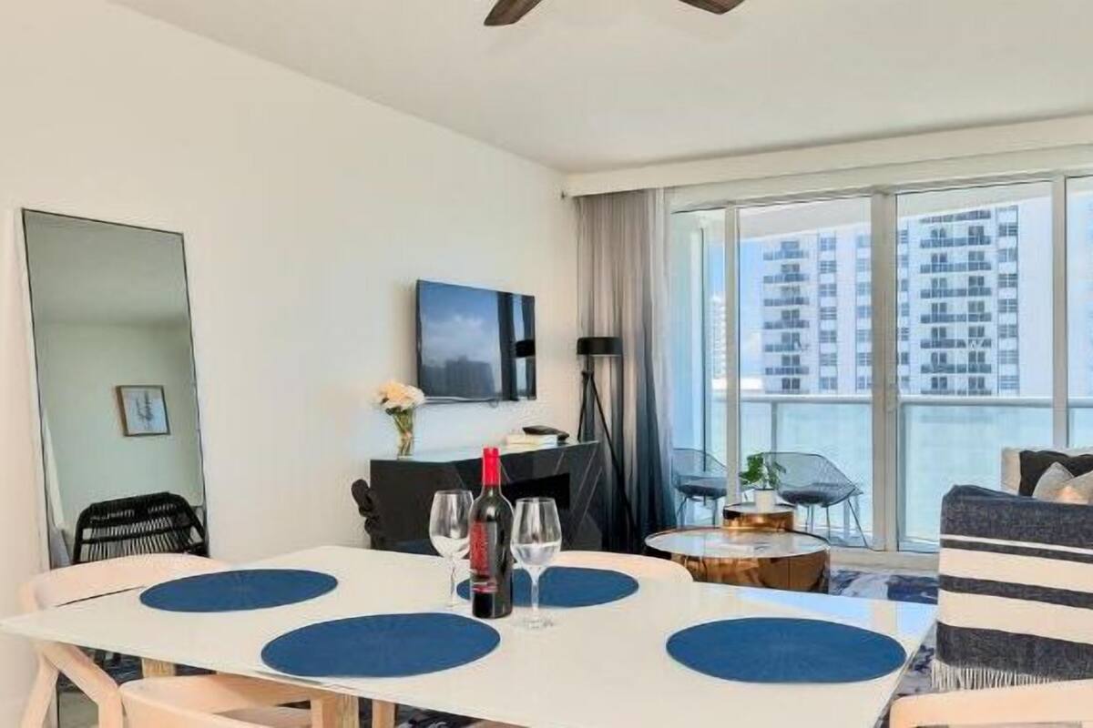 A modern hotel room with a dining area, a sofa, and a large window offering a city view.
