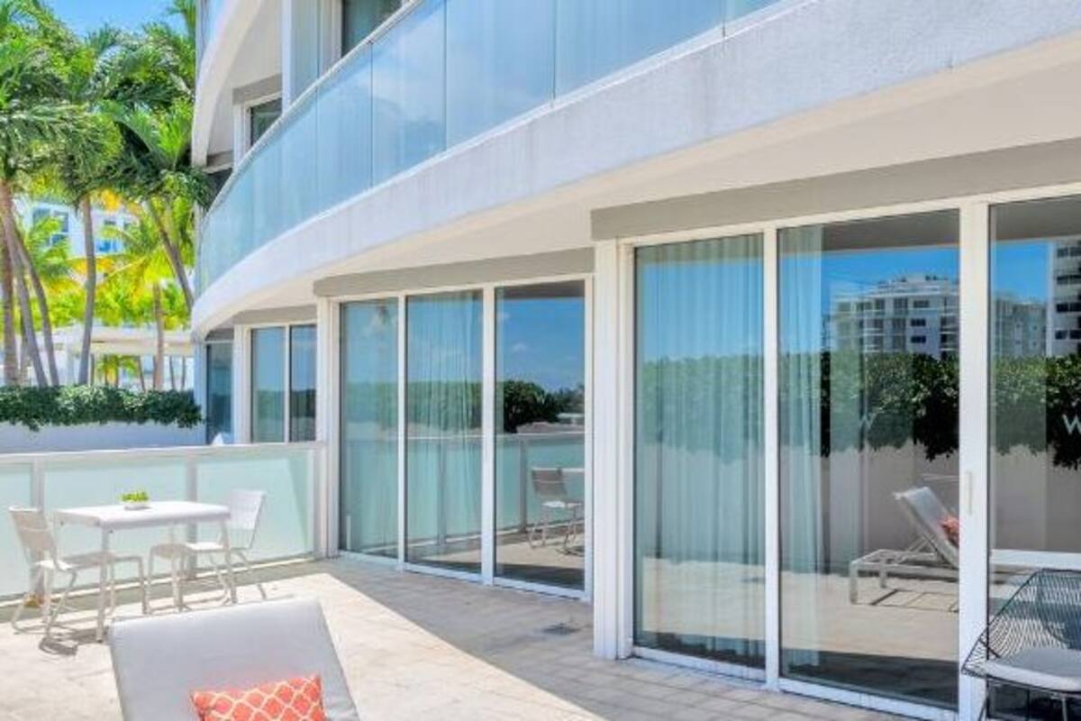 A modern hotel balcony with glass doors, white furniture, and a view of palm trees.