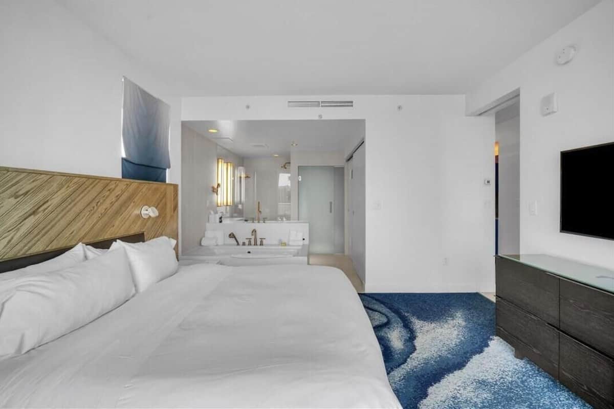 A modern hotel room with a large bed, a wooden headboard, a flat-screen TV, a dark wooden dresser, and a bathroom with a large mirror and white fixtures.