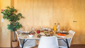 Una mesa de comedor dispuesta con diversos platos y bebidas, rodeada de sillas blancas, en una habitación con paneles de madera y una planta en maceta.