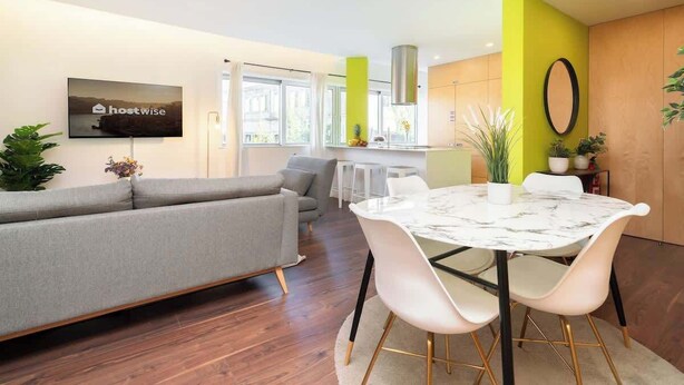 Una sala de estar moderna con un sofá gris, una mesa de comedor blanca con sillas, un televisor de pantalla plana en la pared y una zona de cocina con una pared de acento verde.