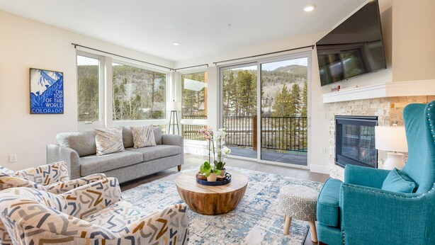A spacious living room with a grey sofa, teal armchair, and patterned ottoman. A wooden coffee table holds decorative items. Large windows offer views of a mountainous landscape. - Brand New Luxury Condo/Mountain Views/Shared Hot Tub (Keystone)