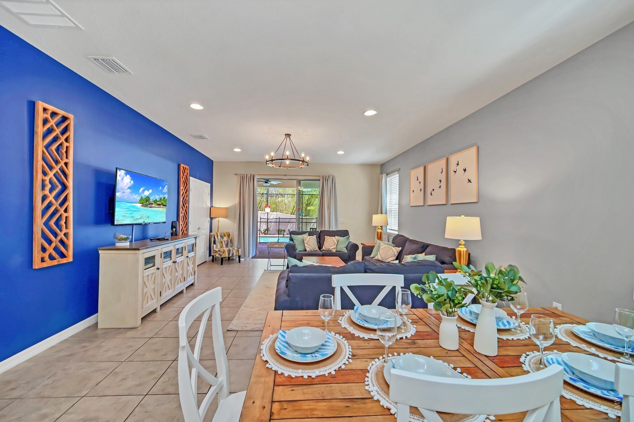 A modern living room with a blue accent wall, a dining area with a wooden table set for two, and a view into the adjacent living space.