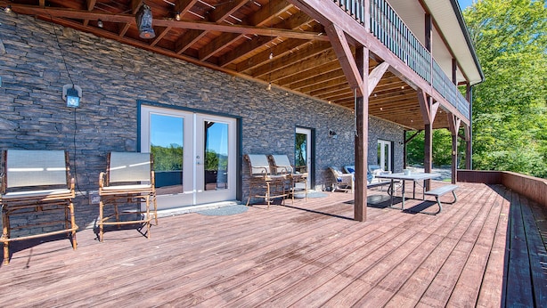 A wooden deck with outdoor furniture, a stone wall, and a covered area with a balcony. - Spacious Smoky Mountain Getaway - Sleeps 10, Game Room, Firepit & Views (Maggie Valley)
